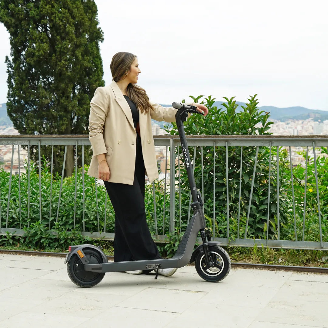 Žena pored crnog električnog romobila Joyor LiteGo na otvorenom s pogledom na grad