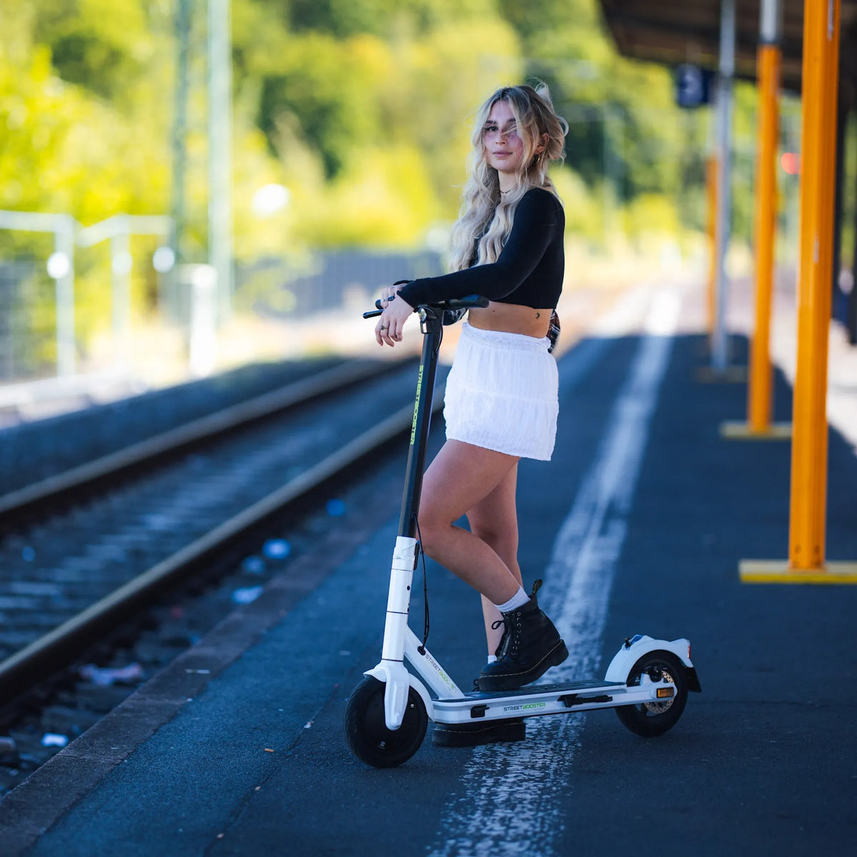 Djevojka stoji na bijelom električnom romobilu Streetbooster Vega na željezničkom peronu