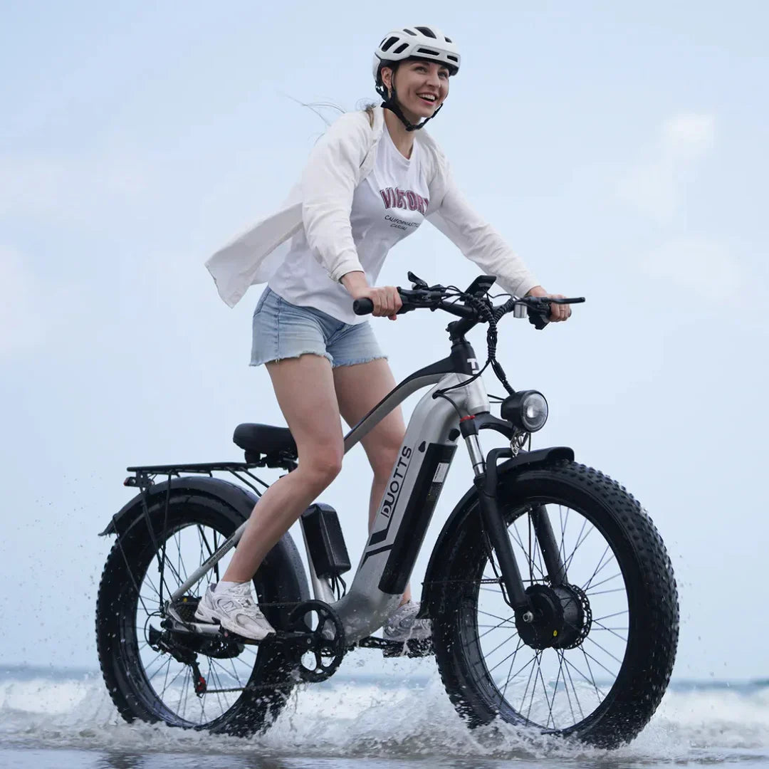 Žena vozi srebrni električni bicikl Duotts F26 kroz plićak na plaži, pogled sa strane