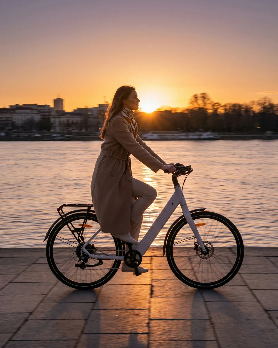 Žena vozi bijeli električni bicikl MS Energy Zorya uz rijeku tijekom zalaska sunca