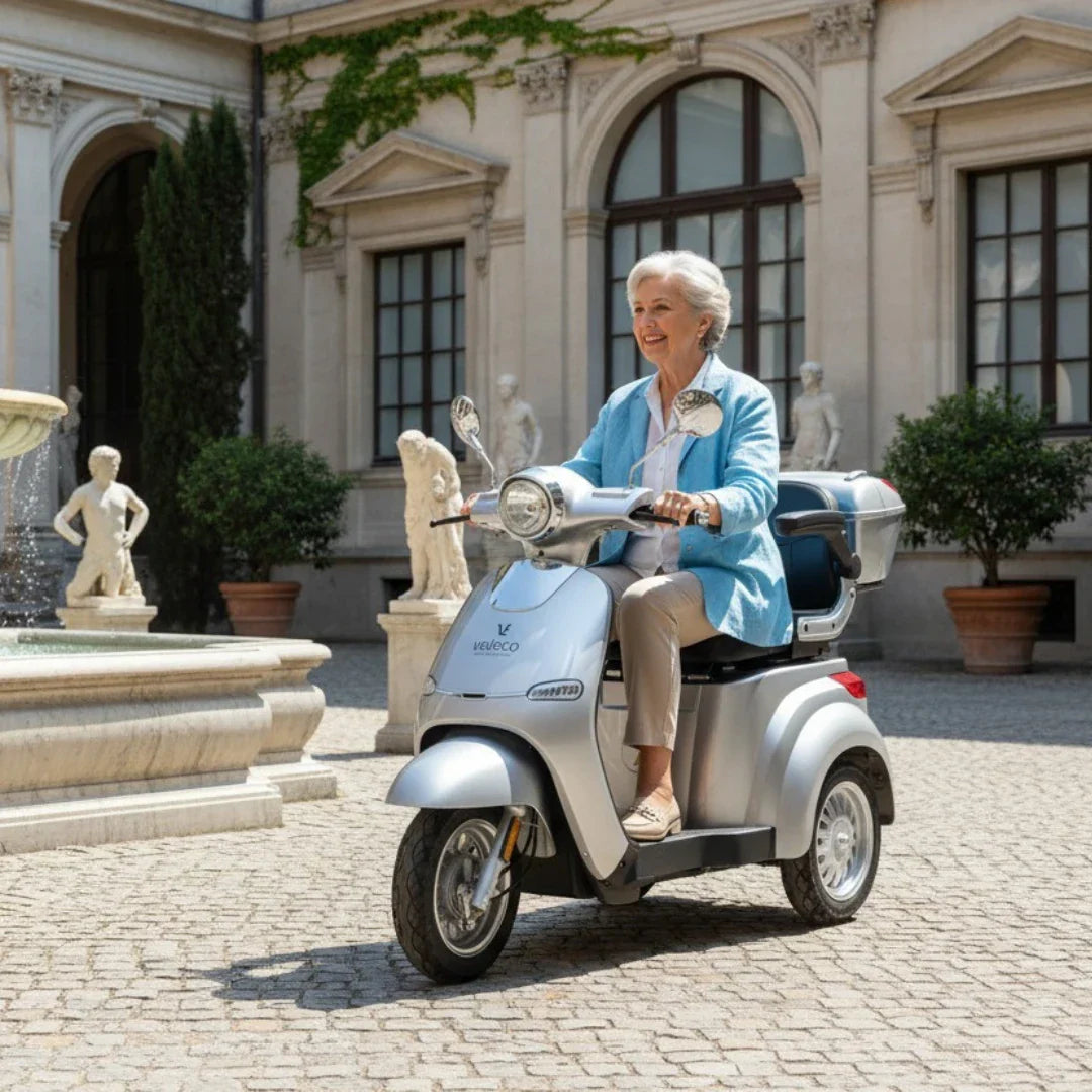 Starija žena vozi srebrni električni skuter za starije osobe Veleco Cristal ispred vile s fontanom