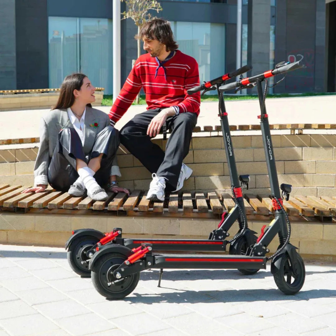 Dva crna električna romobila Joyor G5 s crvenim detaljima pored para na klupi u gradu