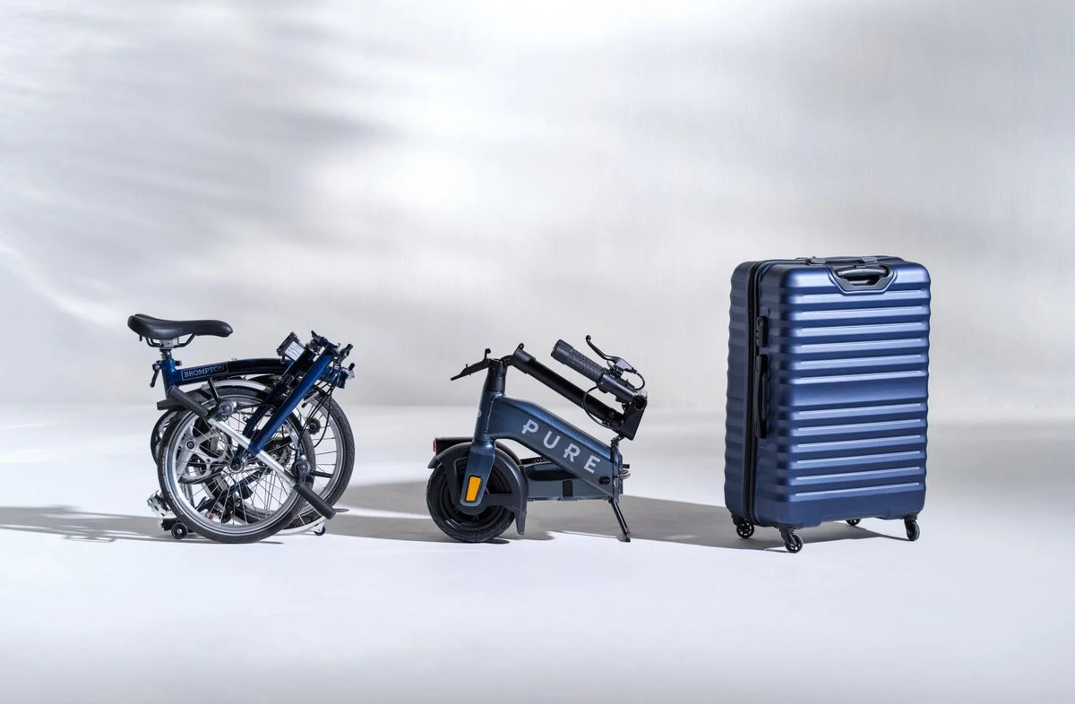 Sklopljeni električni romobil Pure Flex pored sklopljenog bicikla Brompton i plavog kovčega