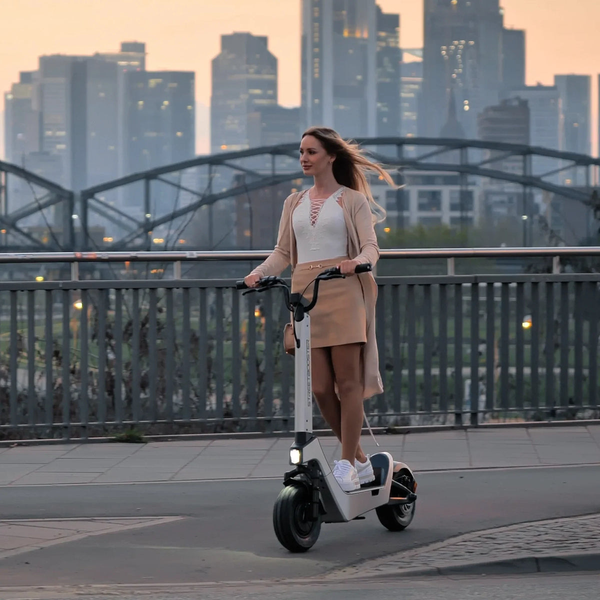 Žena vozi bijeli električni romobil Streetbooster Pollux na gradskom mostu uz pogled na panoramu