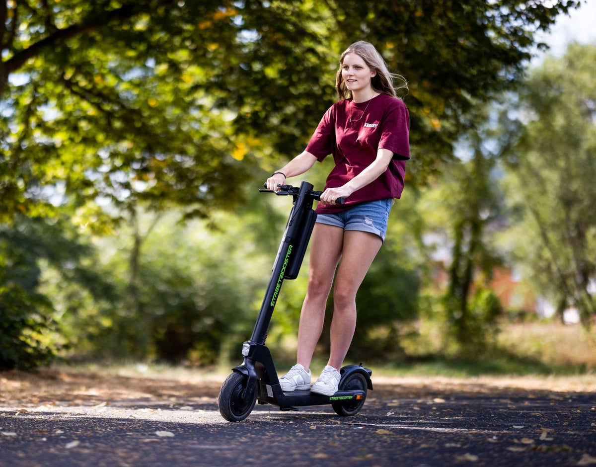 Djevojka vozi crni električni romobil Streetbooster Sirius na asfaltiranoj stazi u prirodi