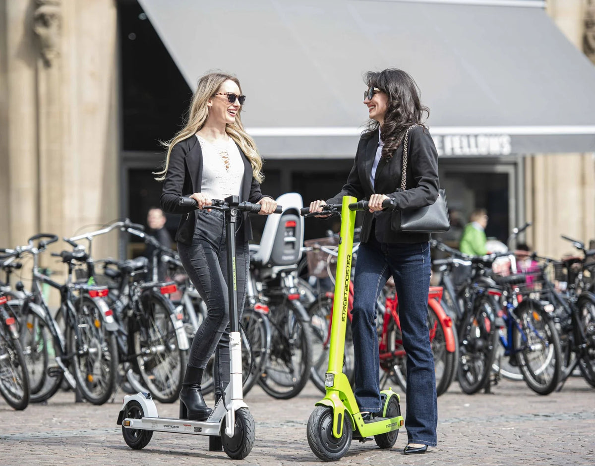 Dvije žene voze bijeli i svijetlozeleni električni romobil Streetbooster Sirius na gradskom trgu