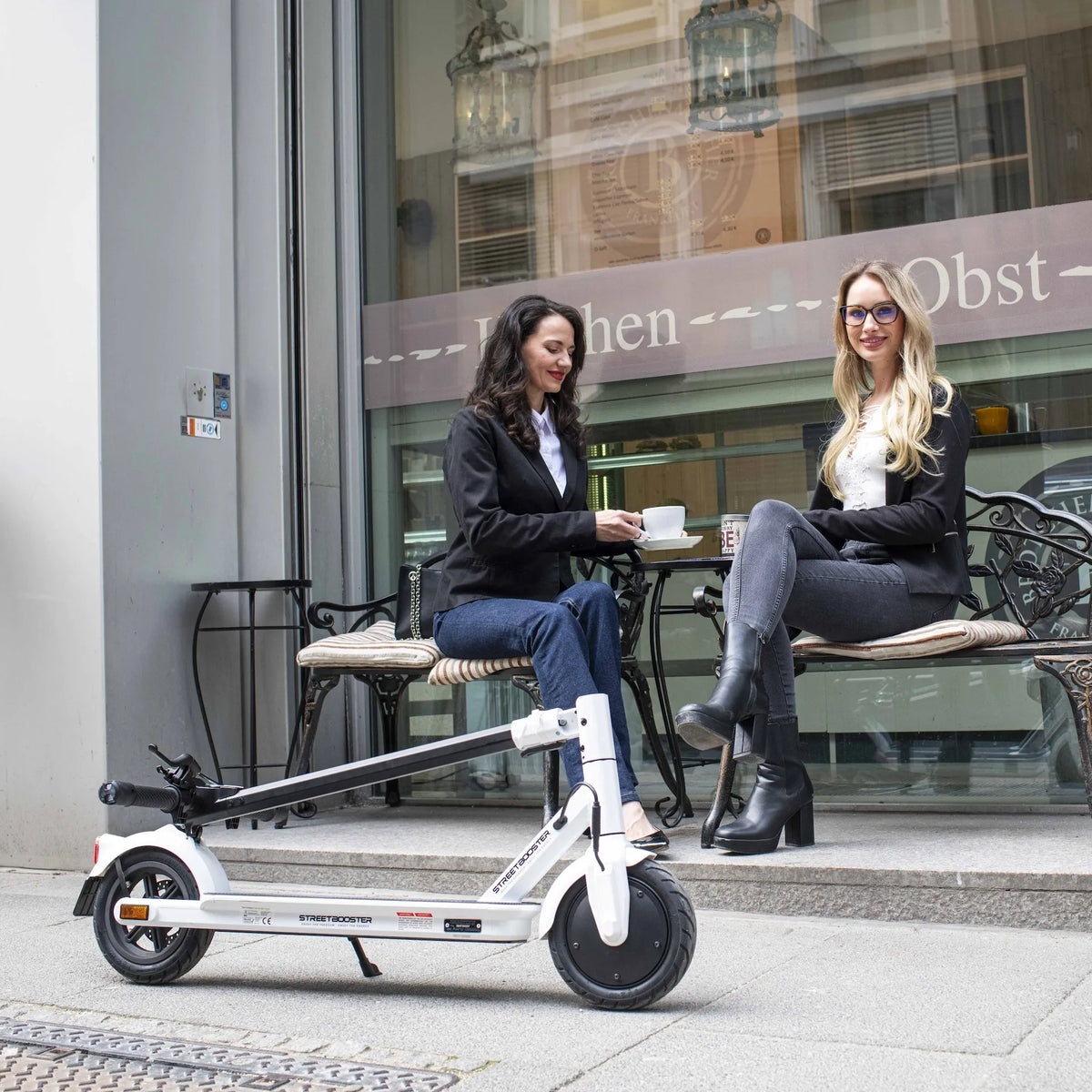Bijeli električni romobil Streetbooster Vega parkiran ispred kafića uz dvije žene u pozadini