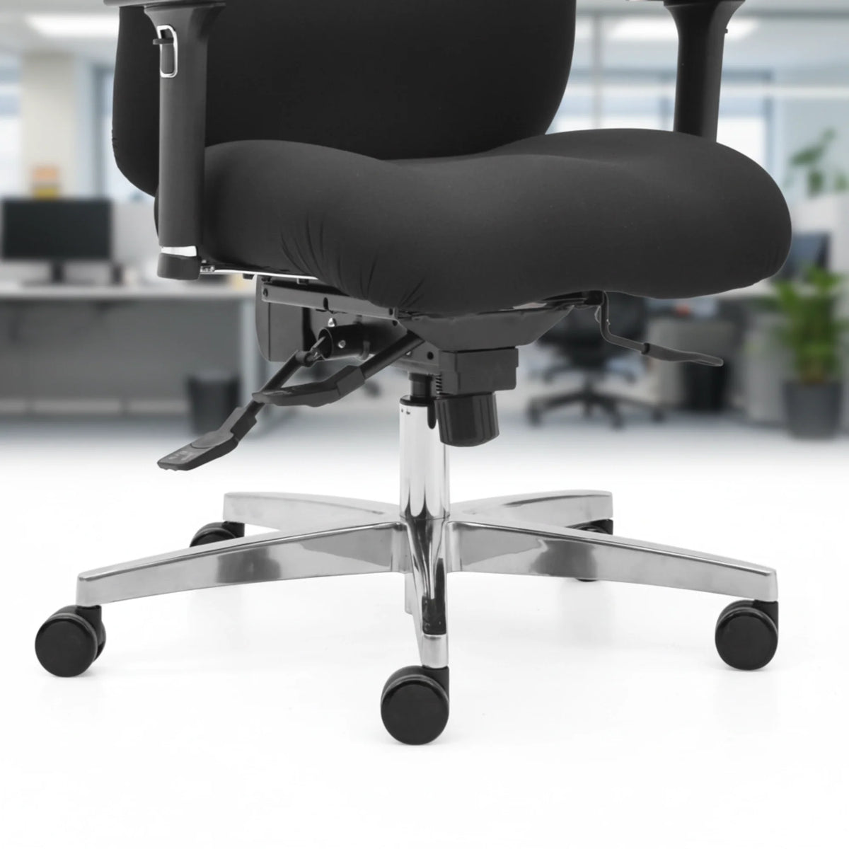 Proizvod je ergonomska uredska stolica, točnije model Hercules. Slika prikazuje krupni plan sjedala i baze stolice, s mekim crnim jastukom, podesivim polugama za visinu i nagib te sjajnom kromiranom bazom s rotirajućim kotačićima. Pozadina sugerira uredsko okruženje s radnim stolovima i biljkama.