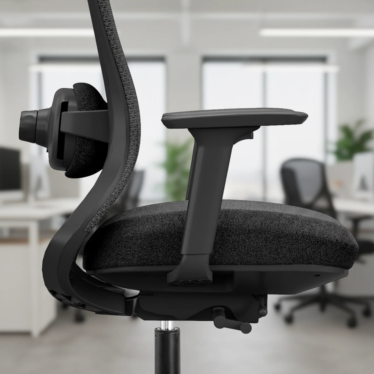 Slika prikazuje ergonomsku uredsku stolicu dizajniranu za udobnost i potporu, vjerojatno model brenda specijaliziranog za uredski namještaj. Stolica ima elegantan dizajn s crnim sjedalom i naslonom od tkanine, podesivim naslonima za ruke i vidljivom funkcijom masaže sa strane. Pozadina sugerira moderno uredsko okruženje s dodatnim stolicama i zelenilom.