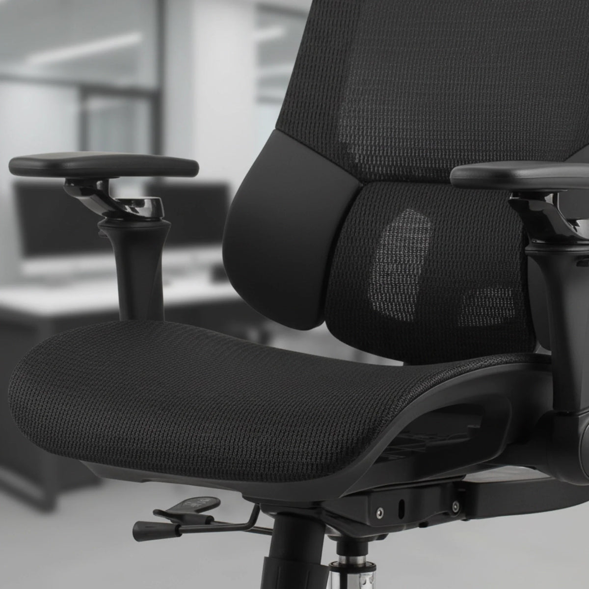 Proizvod je ergonomska uredska stolica, točnije model Trek Plus. Slika prikazuje krupni plan sjedala i naslona za leđa stolice, s dizajnom od crne mrežaste tkanine koji potiče prozračnost. Nasloni za ruke su podesivi, a vidljiva je i poluga za podešavanje visine, što ukazuje na fokus na udobnost i prilagodbu korisniku.