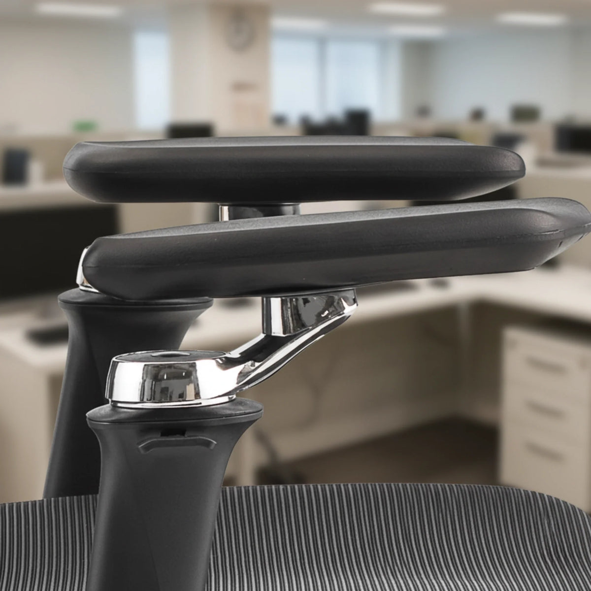 Proizvod je ergonomska uredska stolica, točnije model Trek Plus. Slika prikazuje dizajn naslona za ruku, ističući njegovu podesivu značajku s elegantnim kromiranim mehanizmom i udobnim crnim podloškom. Pozadina sugerira moderno uredsko okruženje s više radnih stanica.